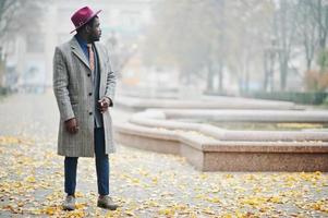 Stylish African American man model in gray coat, jacket tie and red hat posed at foggy weather street. photo