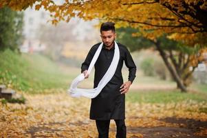 hombre indio con estilo en ropa tradicional negra con bufanda blanca posada al aire libre contra el árbol de hojas amarillas de otoño. foto