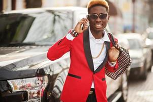 modelo de hombre afroamericano de moda con traje rojo, con cabello destacado contra un gran auto negro y hablando por teléfono móvil. foto