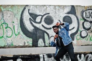 African american man in jeans jacket, beret and eyeglasses against graffiti wall with skull. photo