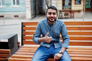 Stylish indian beard model man in casual clothes posed outdoor, sitting on bench at street of India and show thumb up. photo