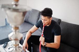 hombre indio guapo y de moda en negro sentado en la habitación, fumando narguile y trabajando en la computadora portátil. foto