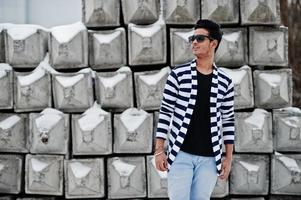 Casual young indian man in stripped sweater and sunglasses posed against stone blocks. photo