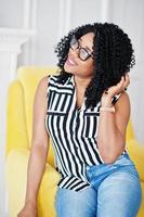 hermosa mujer afroamericana con cabello afro rizado y anteojos posada en la habitación, sentada en una silla amarilla. foto