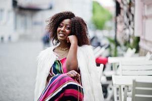 Fashionable african american woman in pink striped jumpsuit with fluffy faux fur coat sitting on chair at outdoor cafe. photo