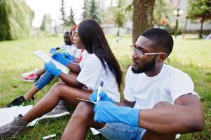 grupo de felices voluntarios africanos sentados bajo un árbol en el parque y escribiendo algo en portapapeles. Concepto de voluntariado, caridad, personas y ecología de África. foto