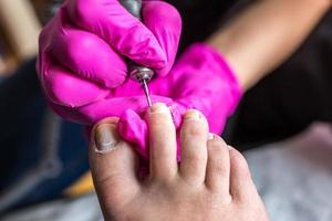 Pedicurist master in pink gloves cuts the cuticle and shellac toe nails in the pedicure salon using drill.  Professional pedicure in cosmetology clinic. Hygiene for feet in beauty salon. photo