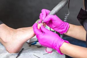 el maestro pedicurista con guantes rosas corta la cutícula y las uñas de los pies de goma laca en el salón de pedicura con un taladro. pedicura profesional en clínica de cosmetología. higiene de los pies en el salón de belleza. foto