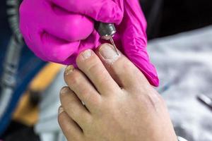 Pedicurist master in pink gloves cuts the cuticle and shellac toe nails in the pedicure salon using drill.  Professional pedicure in cosmetology clinic. Hygiene for feet in beauty salon. photo