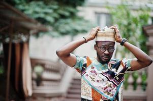 apuesto hombre afroamericano vestido con ropa tradicional, gorra y anteojos en la ciudad moderna. foto