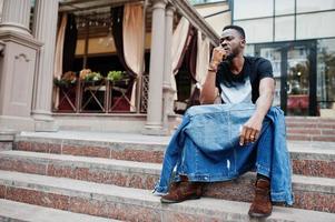 Young attractive serious african american man model. Modern handsome guy in jeans jacket sitting on urban city street. photo