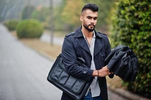 Fashionable tall arab beard man wear on black coat with umbrella and bag case posed at rain weather day. photo