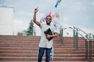 African student male posed with backpack and school items on yard of university, against flags of different countries. Show finger up. photo
