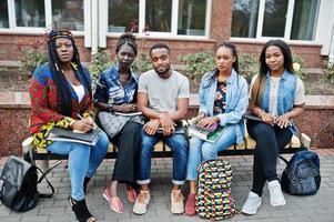 grupo de cinco estudiantes universitarios africanos que pasan tiempo juntos en el campus en el patio de la universidad. amigos afro negros que estudian en un banco con artículos escolares, computadoras portátiles. foto