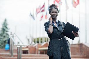 African student female posed with backpack and school items on yard of university, against flags of different countries. photo