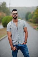 el hombre de barba árabe alto de moda usa camisa, jeans y gafas de sol posados en el parque y sostiene la billetera a mano. foto