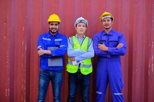 Portrait of three Asian male industry workers with safety uniforms on metal sheet background, arms crossed, looking at camera and smile, cargo shipping, professional freight transportation business. photo