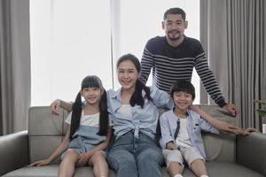 Portrait of Asian Thai family, adult dad, mum with little children happiness home living together, looking at camera, leisure on sofa in white room, lovely weekend and wellbeing domestic lifestyle. photo