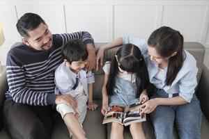 Asian Thai family, adult dad, mum, and children happiness home living relaxing activities and reading book together, leisure on sofa in white room house, lovely weekend, wellbeing domestic lifestyle. photo