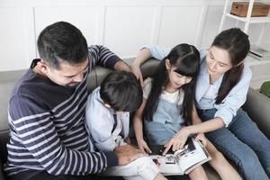 Asian Thai family, adult dad, mum, and children happiness home living relaxing activities and reading book together, leisure on sofa in white room house, lovely weekend, wellbeing domestic lifestyle. photo