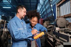 Asian male and female African American engineers in safety uniform work by inspecting machines' voltage current, checking, and maintaining at manufacture factory, electric system service occupations. photo