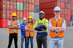 Portrait of a group of multiracial workers team in safety uniforms, arms crossed and looking at camera at logistics dock with many stacks of containers, shipping goods, cargo transportation industry. photo
