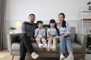 Portrait of Asian Thai family, adult dad, mum with little children happiness home living together, looking at camera, leisure on sofa in white room, lovely weekend and wellbeing domestic lifestyle. photo