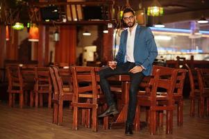 Handsome well-dressed arabian man with glass of whiskey and cigar posed at pub. photo