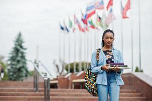 African student female posed with backpack and school items on yard of university, against flags of different countries. photo