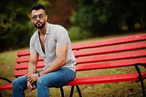 Fashionable tall arab beard man wear on shirt, jeans and sunglasses sitting on red bench at park. photo