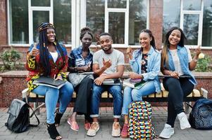 grupo de cinco estudiantes universitarios africanos que pasan tiempo juntos en el campus en el patio de la universidad. amigos afro negros que estudian en un banco con artículos escolares, computadoras portátiles. foto