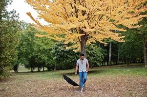 el hombre de barba árabe alto de moda usa camisa, jeans y gafas de sol posados en el parque de otoño con paraguas contra el árbol de hojas amarillas. foto