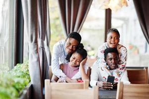 Happy african friends sitting and chatting in cafe. Group of black peoples meeting in restaurant hugs and look at their mobile phone. photo