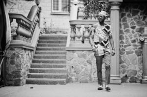 apuesto hombre afroamericano vestido con ropa tradicional, gorra y anteojos en la ciudad moderna. foto