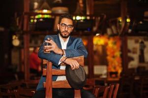 Handsome well-dressed arabian man with glass of whiskey and cigar posed at pub. photo