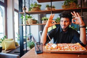 un joven indio confiado con camisa negra sentado en una pizzería con pizza y mostrando el cartel de ok con las manos. foto