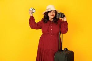 atractiva mujer viajera del sur de asia con vestido rojo intenso, sombrero posado en el estudio sobre fondo amarillo con maleta y cámara de fotos antigua.