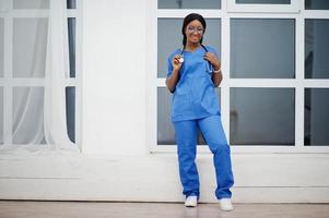 retrato de una joven y feliz doctora afroamericana pediatra con uniforme azul y estetoscopio contra la ventana del hospital. cuidado de la salud, médico, especialista en medicina - concepto. foto