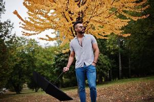 Fashionable tall arab beard man wear on shirt, jeans and sunglasses posed on autumn park with umbrella against yellow leaves tree. photo