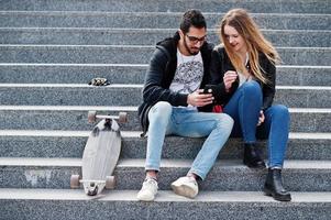 Cool multiracial couple sitting on stairs with longboard and look in mobile phone. photo