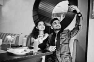 Lovely indian couple in love, wear at saree and elegant suit, sitting on restaurant and making selfie by phone together. On table dessert cakes, ice cream and juice. photo