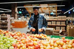 elegante hombre afroamericano casual con chaqueta de jeans y boina negra revisando frutas de manzana en la sección orgánica del supermercado.