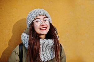 retrato de una chica morena con bufanda gris y sombrero, gafas en clima frío con sol contra la pared naranja de la casa antigua. espacio de copia libre. foto
