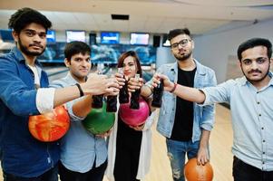 Group of five south asian peoples having rest and fun at bowling club. Clinking cold soda drinks from glass bottles and bowling balls at hands. photo