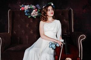 Pretty young gilrl musician in white dress with double bass sitting on brown vintage sofa. photo