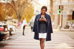 Success stylish african american woman in coat walking at crosswalk and looking on phone. photo