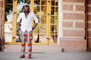 Stylish african american man in white shirt and colored pants with hat and glasses posed outdoor. Black fashionable model boy. photo