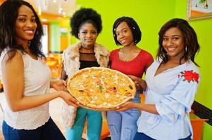 Four young african girls in bright colored restaurant hold wooden tray with pizza in hands. photo