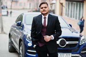 Rich indian businessman in formal wear standing against business suv car. photo