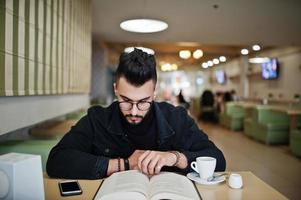 el hombre árabe usa una chaqueta de jeans negros y anteojos sentados en un café, lee un libro y bebe café. chico modelo árabe elegante y de moda. foto